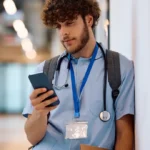 Junger Arzt lehnt nachdenklich mit Smartphone und Unterlagen unter dem Arm an einer Wand