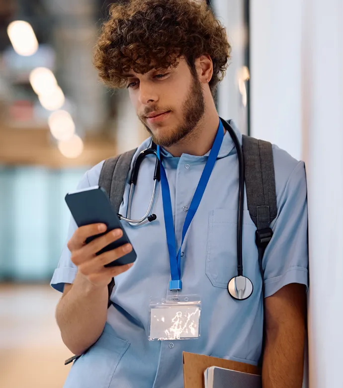 Junger Arzt lehnt nachdenklich mit Smartphone und Unterlagen unter dem Arm an einer Wand
