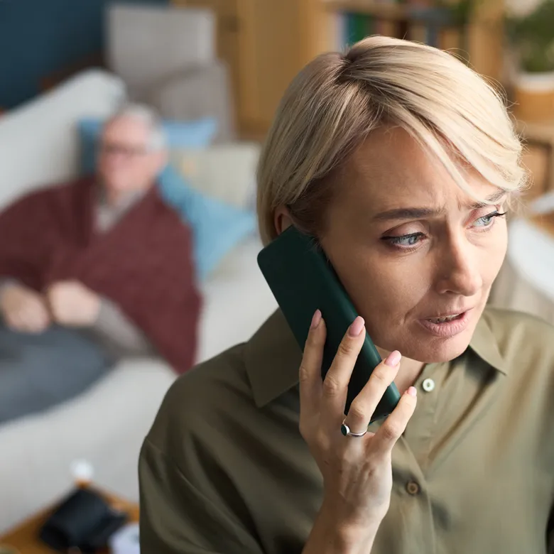 Frau telefoniert mit besorgtem Gesichtsausdruck, ein Patient ruht im Hintergrund auf einem Sofa